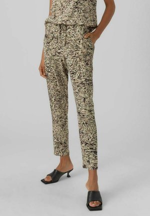 Vrouw draagt beige broek met abstract zwart en bruin patroon, gecombineerd met zwarte open-toe pumps met hak, staand met één hand in de zak.