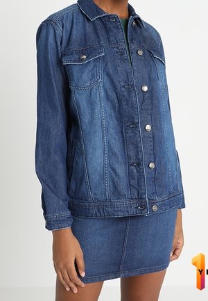 Vrouw die een middelblauwe spijkerjasje met knopen en borstzakken draagt, samen met een bijpassende korte spijkerrok.