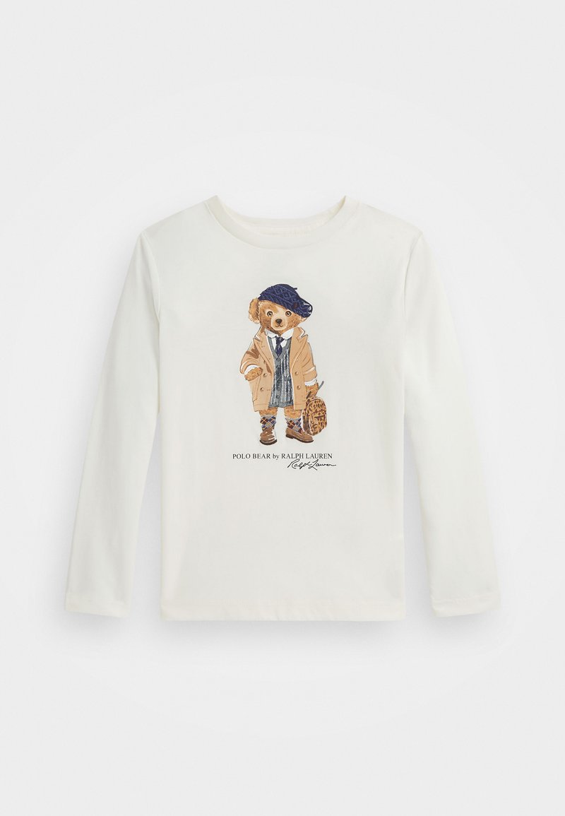 Biela košeľa s dlhým rukávom, ktorá zobrazuje grafiku medveďa v béžovom kabáte a modrom barete, ktorý drží tašku. Text pod obrázkom znie: "Polo Bear od Ralph Lauren."