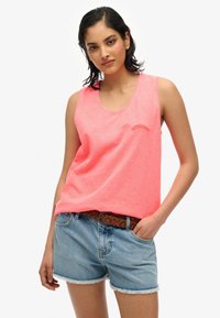 Różowy bezrękawnik z okrągłym dekoltem i subtelnym logo. W połączeniu z jasnoniebieskimi dżinsowymi szortami z postrzępionymi brzegami i plecionym brązowym paskiem.