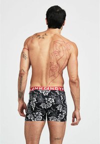 Svarta boxerkalsonger med vita ros- och spindelnätsmönster, röd märkestub, modell med tatueringar synliga på ryggen.