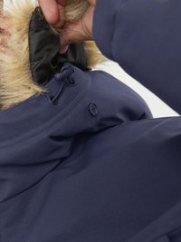 Giacca blu scuro con cappuccio foderato in pelliccia sintetica. Le caratteristiche includono un cordoncino, chiusura a bottoni e una texture del tessuto liscia. Vista ravvicinata.
