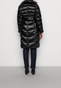 Abrigo negro acolchado con un patrón enChevron, cuello alto y cintura con cinturón. Lleva puesto unos jeans oscuros y botas hasta el tobillo. Textura suave y brillante.