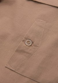 Gros plan sur un tissu beige avec un bouton blanc cousu sur une poche rectangulaire avec des coutures visibles.