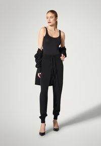 Sort t ætsiddende bodysuit, matchende joggingbukser med ankelmanchetter og en løs sort jakke hængende over skuldrene. Høje hæle fremhæver outfittet.