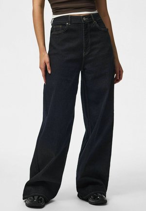 Personne portant un jean bleu foncé à jambes larges et des chaussures noires, les mains détendues le long du corps sur un fond uni.