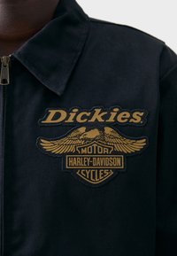 Black fabric jacket with a zipper, featuring an embroidered gold and black logo that includes wings and text: "Dickies Motor Harley-Davidson Cycles."