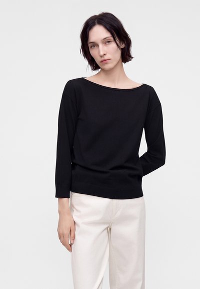 Jeune femme aux cheveux courts et foncés, vêtue d'un haut noir à manches longues et d'un pantalon blanc, debout devant un fond blanc uni.