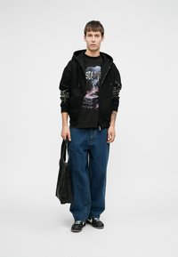 Črna zip-up kapuca z vzorčastimi rokavi preko grafične črne majice. Ohlapne modre kavbojke in črne superge. Drži črno platneno torbo.