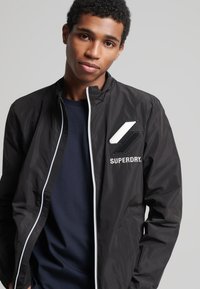 Chaqueta bomber ligera negra con cremallera blanca y logo bordado. Cuenta con un cuello alzado y una textura suave y brillante.