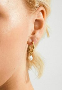 Guld hoop ørering med en hængende hvid perle, båret på venstre øre, sat mod lys hud og kort blond hår.