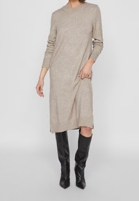 Vestito in maglia beige con maniche lunghe e orlo a coste, caratterizzato da spacchi laterali nella parte inferiore. Abbinato a stivali neri in pelle al ginocchio.