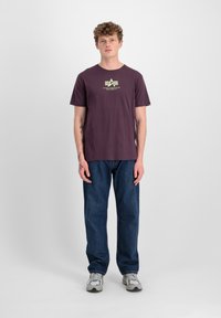 Tmavě červené bavlněné tričko s grafickým logem, kombinované s modrými džíny a šedými sportovními botami. Jednoduchý design, normální střih.