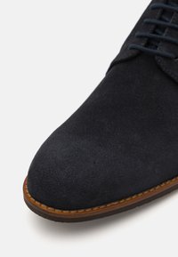 Scarpa in suede blu navy con punta rotonda, presenta lacci scuri e una suola strutturata marrone. Superficie superiore liscia e delicate cuciture.