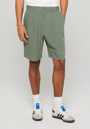 Homme portant un short vert clair ajusté avec des plis devant, des chaussettes blanches et des baskets blanches avec des accents bleus et noirs sur un fond gris.