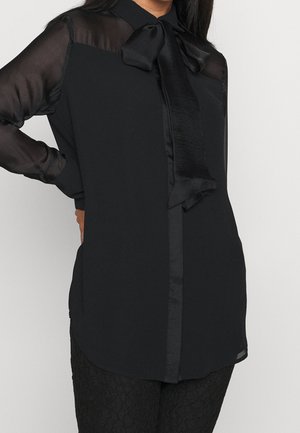 Kvinde iført sort, gennemskinnelig bluse med lange ærmer og satin sløjfe ved kraven samt matchende sorte, teksturerede bukser.