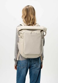 Béžový batoh s rolovacím vrchem, nastavitelnými popruhy, bočními kapsami a přední kapsou na zip; vyrobený z hladkého, voděodolného materiálu.