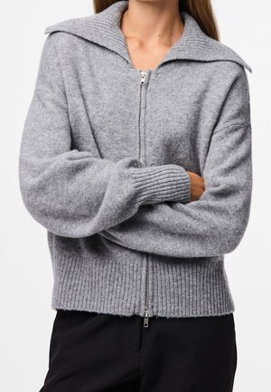 Femme portant un pull en maille gris à fermeture éclair avec un large col et des poignets et ourlet côtelés, bras croisés, assorti à un pantalon noir.