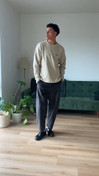 Suéter texturizado bege claro, calças soltas cinza escuro e sapatos pretos. Ambiente interior com sofá verde e plantas ao fundo.