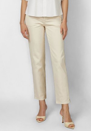 Woman wearing beige straight-leg pants and matching high-heeled open-toe shoes, standing against a plain light background.