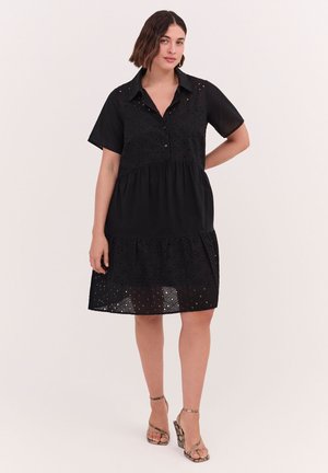 Femme portant une robe noire à manches courtes avec des détails en œillets et des sandales à talons hauts en peau de serpent, debout devant un fond uni.