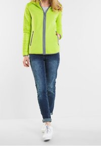 Světlá zelená zipová bunda s šedými detaily, která má kapuci a dvě přední kapsy, doplněná tmavými džíny a bílými teniskami.