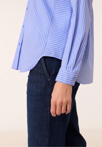 Camicia a righe blu chiaro con orlo curvo, maniche lunghe e polsini con bottoni, abbinata a jeans in denim scuro e un dettaglio con bottone visibile.