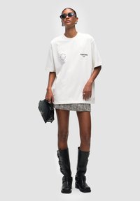 T-shirt bianco oversize con collo rotondo, design grafico, abbinato a una mini gonna a fantasia, stivali neri al ginocchio e occhiali da sole eleganti.