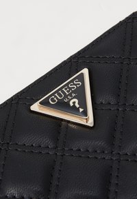 Schwarze gesteppte Handtasche mit einem dreieckigen, schwarzen und goldenen Guess-Logo-Anhänger mit einem Fragezeichen in der Mitte. Hergestellt aus strukturiertem synthetischem Material.