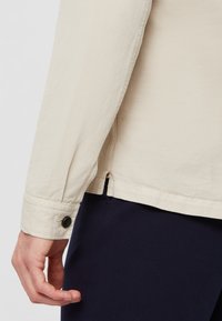 Camicia in cotone beige con maniche lunghe, chiusura con bottoni sui polsini, una fessura laterale e una texture liscia. Abbinata a pantaloni blu scuro.