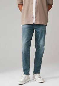 Jeans din denim albastru deschis cu croială dreaptă, având o ușoară decolorare și hardware minim. Asortate cu o cămașă bej cu mânecă scurtă și teniși albi.