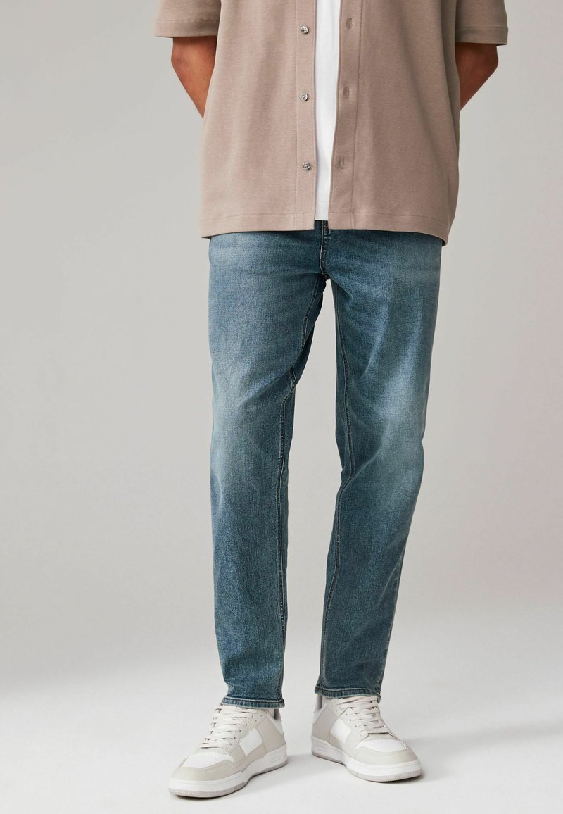 Jeans de mezclilla azul claro con corte recto, con un desvanecimiento sutil y hardware mínimo. Combinados con una camiseta de manga corta beige y zapatillas blancas.