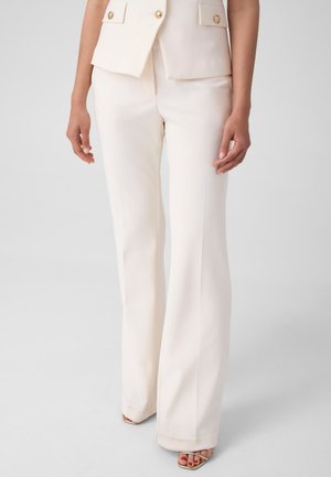 Femme portant un pantalon blanc à larges jambes associé à un gilet blanc assorti avec des boutons dorés et des sandales pieds nus avec du vernis à ongles rouge.