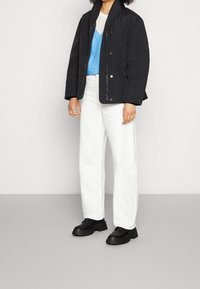 Svart vadderad jacka med ståkrage, som bärs tillsammans med en blå skjorta och vita vida byxor. Svarta klumpiga skor fulländar outfiten.