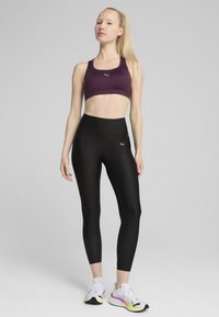 Lila sportmelltartó, fekete, magas derekú leggings, mindkettő sima és testhezálló. Fehér sneaker, multicolor részletekkel kiegészíti az öltözetet.