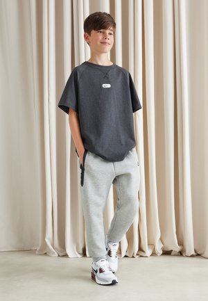 Ragazzo che indossa una t-shirt Nike grigio scuro, pantaloni della tuta grigio chiaro e sneakers Nike bianche, in piedi con le mani nelle tasche davanti a tende beige.