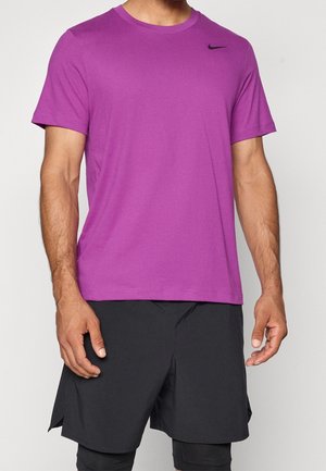 Homme portant un t-shirt Nike violet à manches courtes et un short de sport noir, debout devant un fond uni.