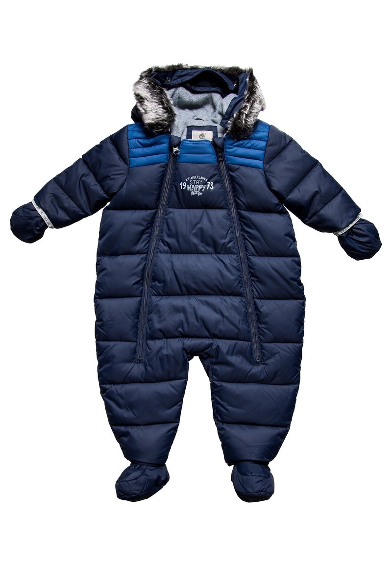 Mörkblå puffy sn overall med en huva med fuskpäls, blixtlås framtill och elastiska muddar. Har quiltat mönster och broderad text på bröstet.