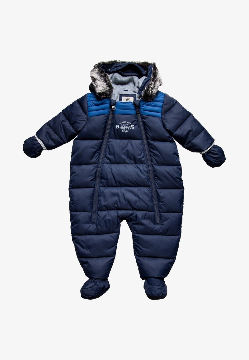 Mörkblå puffy sn overall med en huva med fuskpäls, blixtlås framtill och elastiska muddar. Har quiltat mönster och broderad text på bröstet.