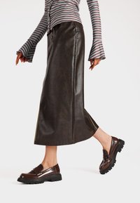 Gonna marrone in faux pelle con spacco laterale, abbinata a mocassini marroni lucidi con suola nera spessa. Camicia a righe con maniche a campana.