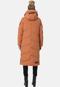 Dikke oranje lange jas met zigzagstiksel, een capuchon en een labeldetail nabij de zoom. gedragen over zwarte, getailleerde broeken en laarzen.