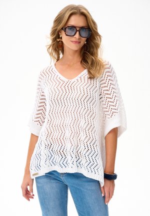 Kobieta w dużych niebieskich okularach przeciwsłonecznych, wiszących kolczykach, białym luźnym swetrze i niebieskich dżinsach, pozująca z jedną ręką swobodnie opuszczoną wzdłuż ciała.