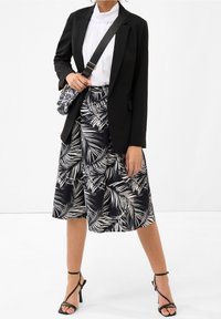 Černý blazer přes bílou blůzu s volány, v kombinaci s černou a bílou sukní se vzorem palmových listů a černými sandály na podpatku.