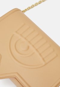 Béžová crossbody taška s texturovaným designem oka, vyrobená z hladké kůže a se zlatým řetízkem. Elegantní a minimalistický tvar se švy.