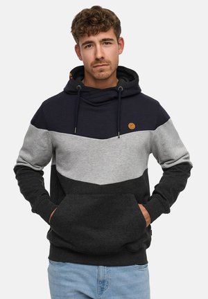 Junger Mann mit lockigem Haar, trägt einen farblich abgesetzten Hoodie in Marineblau, Grau und Schwarz mit den Händen in der vorderen Tasche, hellblaue Jeans.