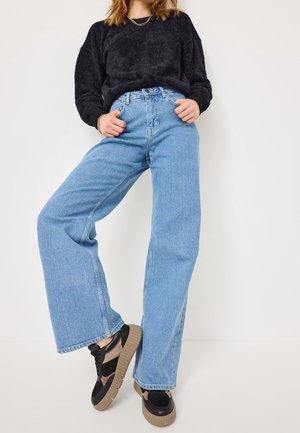 Persona che indossa jeans a gamba larga di colore blu chiaro, un maglione nero in pelliccia sintetica, scarpe da ginnastica con plateau nere e beige, in piedi contro un muro bianco.