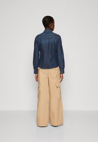 Denim srajca v temno modri barvi z dolgimi rokavi in gumbi na manšeti, v kombinaciji s svetlo bež cargo hlačami, ki imajo stranske žepe in sproščen kroj.