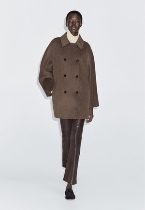 Brauner Wollmantel mit doppelter Knopfleiste, großem Kragen und schwarzen Knöpfen. Kombiniert mit enganliegenden braunen Lederhosen. Glatte Textur.