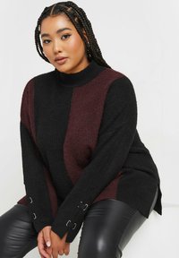 Överdimensionerad stickad tröja i svart och burgunderröd med hög halsringning, långa ärmar och dekorativa spännen vid ärmsluten. Slät textur.