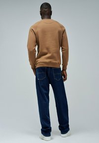Sudadera marrón con puños y dobladillo acanalados, combinada con jeans de mezclilla azul oscuro de corte holgado. El conjunto se complementa con zapatillas blancas.